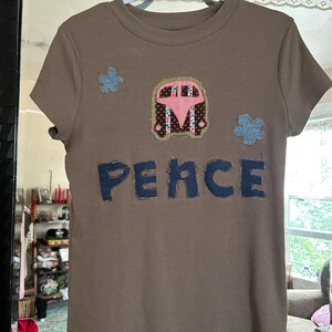 Könnte beinhalten: Ein braunes Kurzarm-T-Shirt mit einer zentralen Applikation eines rosa-braunen Vans, zwei blauen blumenförmigen Aufnähern und dem Wort "PEACE" in großen, dunkelblauen Stoffbuchstaben mit sichtbaren Nähten im Brustbereich.