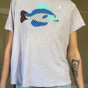May include: A light purple short-sleeved t-shirt featuring a large fabric appliqué of a fish. The fish is constructed from blue, teal, and white patterned fabric pieces with a brown tail and a small red button eye. Three white buttons are sewn above the fish.