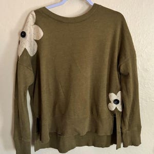 Könnte beinhalten: Ein olivgrünes Langarm-Sweatshirt mit Rundhalsausschnitt und gerippten Bündchen. Das Kleidungsstück ist mit zwei großen, cremefarbenen Stoffblumen-Applikationen verziert, die jeweils mit einem schwarzen Knopf an der rechten Schulter und linken Seite.