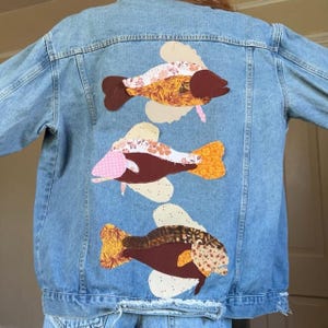 Op de afbeelding: Een lichtblauw spijkerjack met drie geappliqueerde visontwerpen op de achterkant. Elke vis is opgebouwd uit verschillende stoffen lapjes in bruin-, oranje- en crèmetinten met bloem- en geometrische patronen, verticaal gerangschikt.