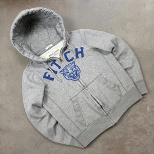 May include: A light grey zip-up hoodie featuring a quilted hood lining and a front zip closure. The chest displays the blue embroidered text FITCH above a blue tiger head graphic. The garment includes two front pockets and ribbed cuffs.