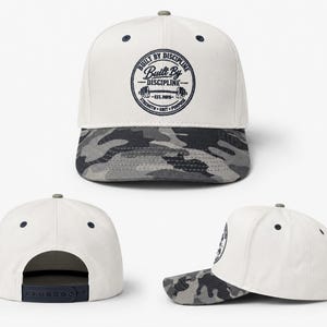 Peut inclure: Une casquette de baseball blanche avec une visière camouflage gris foncé et une fermeture snapback bleu marine. L'avant présente un logo circulaire avec le texte "Built By Discipline" et "Est. 2026" autour d'un haltère et "Strength Grit Purpose".