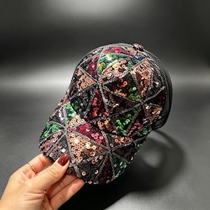 Puede incluir: Una gorra de béisbol con un patrón geométrico de triángulos cubiertos de lentejuelas multicolores. Las lentejuelas muestran tonos rosa, verde y negro, separados por costuras oscuras. La parte trasera de la gorra está hecha de malla negra.