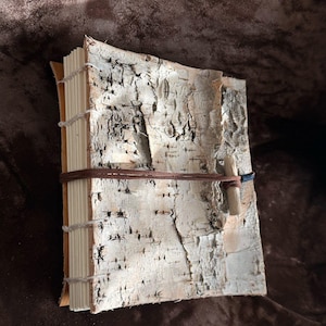 May include: A handmade journal featuring a cover made from textured white and grey birch bark. The pages are bound with visible light-colored thread along the spine. A thin brown leather cord wraps around the cover, secured by a small wooden toggle.