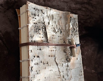 Puede incluir: Un diario hecho a mano con una cubierta de corteza de abedul texturizada en blanco y gris. Las páginas están encuadernadas con hilo claro visible a lo largo del lomo. Un cordón fino de cuero marrón envuelve la cubierta, asegurado por un pequeño pasador de madera.