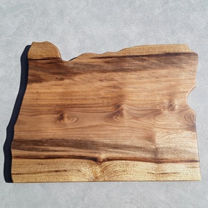 May include: A wooden cutting board shaped like the state of Oregon. The board has a natural wood finish and a dark brown edge.