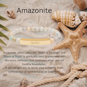 May include: A beaded bracelet featuring smooth, round, multi-coloured amazonite beads in shades of pale blue, cream, and soft grey. The bracelet includes a central, polished gold-tone metal bar engraved with the word Goddess in script font.