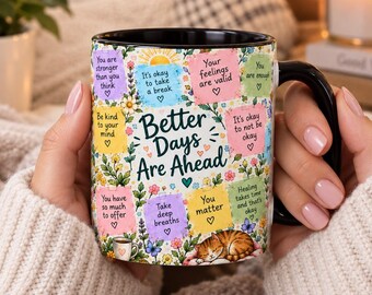 May include: A black ceramic mug featuring a white background with colorful pastel squares containing positive affirmations like Better Days Are Ahead, You matter, and It's okay to take a break, decorated with floral patterns and a sleeping cat.