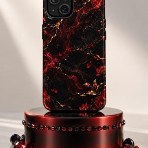 May include: A smartphone case featuring a black and red marble pattern with gold veining and small red floral accents. The case is displayed on a cylindrical stand decorated with a row of red and black crystals and surrounded by loose red and black gems.