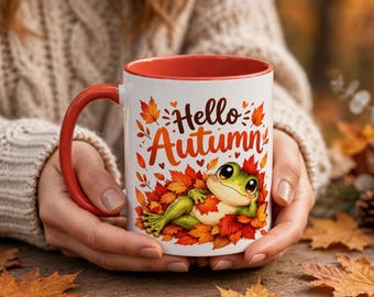 May include: A white ceramic mug with a red interior and handle, featuring a graphic of a green frog resting in a pile of orange and red autumn leaves. The text "Hello Autumn" is printed in a brown script font above the frog illustration.
