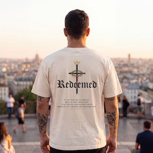Op de afbeelding: Een cr&egrave;mekleurig T-shirt met korte mouwen en een afbeelding op de achterkant. Het ontwerp bevat een kruis met een doornenkroon, het woord "Redeemed" in zwarte gotische letters en de tekst "If any man be in Christ, he is a new creature."