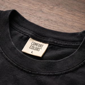 Op de afbeelding: Een zwart t-shirt met een geribbelde ronde hals en een cr&egrave;mekleurig stoffen label in de nek. Het label bevat de zwarte tekst COMFORT COLORS en de letter L, die de maat aangeeft, gedrukt in een duidelijk, schreefloos lettertype op het kledingstuk.