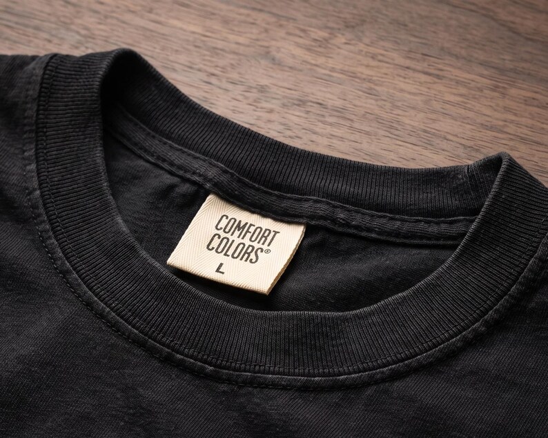 Op de afbeelding: Een zwart t-shirt met een geribbelde ronde hals en een cr&egrave;mekleurig stoffen label in de nek. Het label bevat de zwarte tekst COMFORT COLORS en de letter L, die de maat aangeeft, gedrukt in een duidelijk, schreefloos lettertype op het kledingstuk.