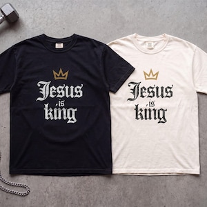 Op de afbeelding: Twee T-shirts met korte mouwen en ronde hals, &eacute;&eacute;n zwart en &eacute;&eacute;n gebroken wit, beide voorzien van een gouden kroonafbeelding boven de verweerde witte tekst Jesus is king. De shirts liggen naast een metalen halter en een zilveren ketting.