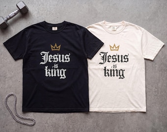 May include: Two short-sleeve crew neck t-shirts, one black and one off-white, each featuring a gold crown graphic above the distressed white text that reads Jesus is king. The shirts are displayed alongside a metal dumbbell and a silver chain.