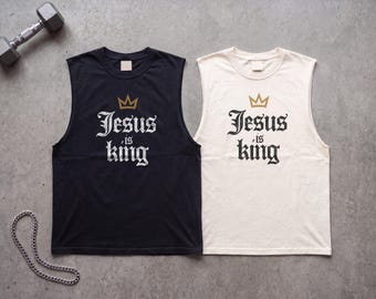 Op de afbeelding: Twee mouwloze shirts, één zwart en één gebroken wit, elk voorzien van een gouden kroonafbeelding boven de tekst "Jesus is king" in een zwart of wit gotisch lettertype. De shirts hebben een ronde hals en liggen op een grijze ondergrond.