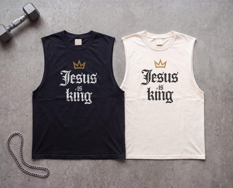 May include: Two sleeveless shirts, one black and one off-white, each featuring a gold crown graphic above the text "Jesus is king" in a black or white gothic-style font. The shirts have a crew neckline and are displayed on a grey surface.