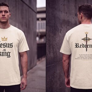Op de afbeelding: Een cr&egrave;mekleurig T-shirt met korte mouwen. De voorkant heeft een gouden kroonicoon boven de zwarte tekst "Jesus is king". De achterkant toont een kruis met een kroon, het woord "Redeemed" en het vers 2 Corinthians 5:17 in zwarte tekst.