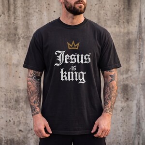 Op de afbeelding: Een zwart t-shirt met korte mouwen voorzien van een gouden kroongrafiek boven de witte tekst in verweerde stijl die Jesus is king leest. Het shirt heeft een ronde hals en een standaard pasvorm, met een opvallende religieuze boodschap op de borst.