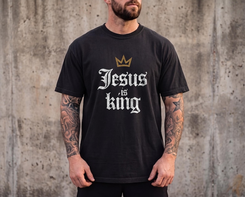 Op de afbeelding: Een zwart t-shirt met korte mouwen voorzien van een gouden kroongrafiek boven de witte tekst in verweerde stijl die Jesus is king leest. Het shirt heeft een ronde hals en een standaard pasvorm, met een opvallende religieuze boodschap op de borst.