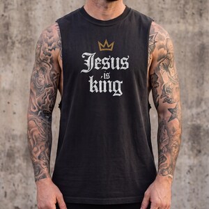 May include: A black sleeveless shirt featuring a gold crown graphic above the white text that reads Jesus is king. The shirt has a crew neckline and wide arm openings, constructed from a solid black fabric with a distressed white font style.