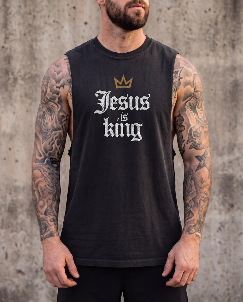 May include: A black sleeveless shirt featuring a gold crown graphic above the white text that reads Jesus is king. The shirt has a crew neckline and wide arm openings, constructed from a solid black fabric with a distressed white font style.