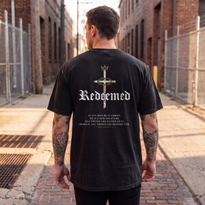 Op de afbeelding: Een zwart t-shirt met korte mouwen en een afbeelding op de achterkant met een kroon, een kruis en een doornenkroon. De tekst luidt "Redeemed" en "If any man be in Christ, he is a new creature. Old things are passed away; behold, all things are become new. 2 Corinthians 5:17."