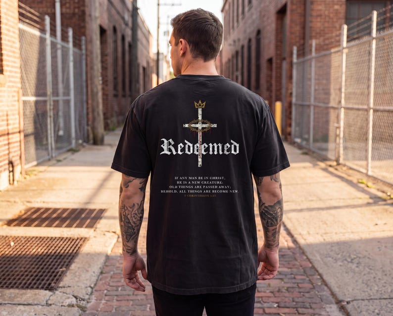 Op de afbeelding: Een zwart t-shirt met korte mouwen en een afbeelding op de achterkant met een kroon, een kruis en een doornenkroon. De tekst luidt "Redeemed" en "If any man be in Christ, he is a new creature. Old things are passed away; behold, all things are become new. 2 Corinthians 5:17."