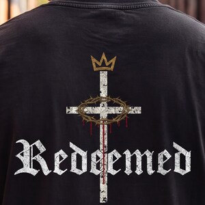 Op de afbeelding: Een zwart t-shirt met een afbeelding op de achterkant. Het ontwerp bevat een wit, verweerd kruis met een gouden kroon erboven, een bruine doornenkroon rond het kruis, rode druppels en het woord "Redeemed" in een wit gotisch lettertype.