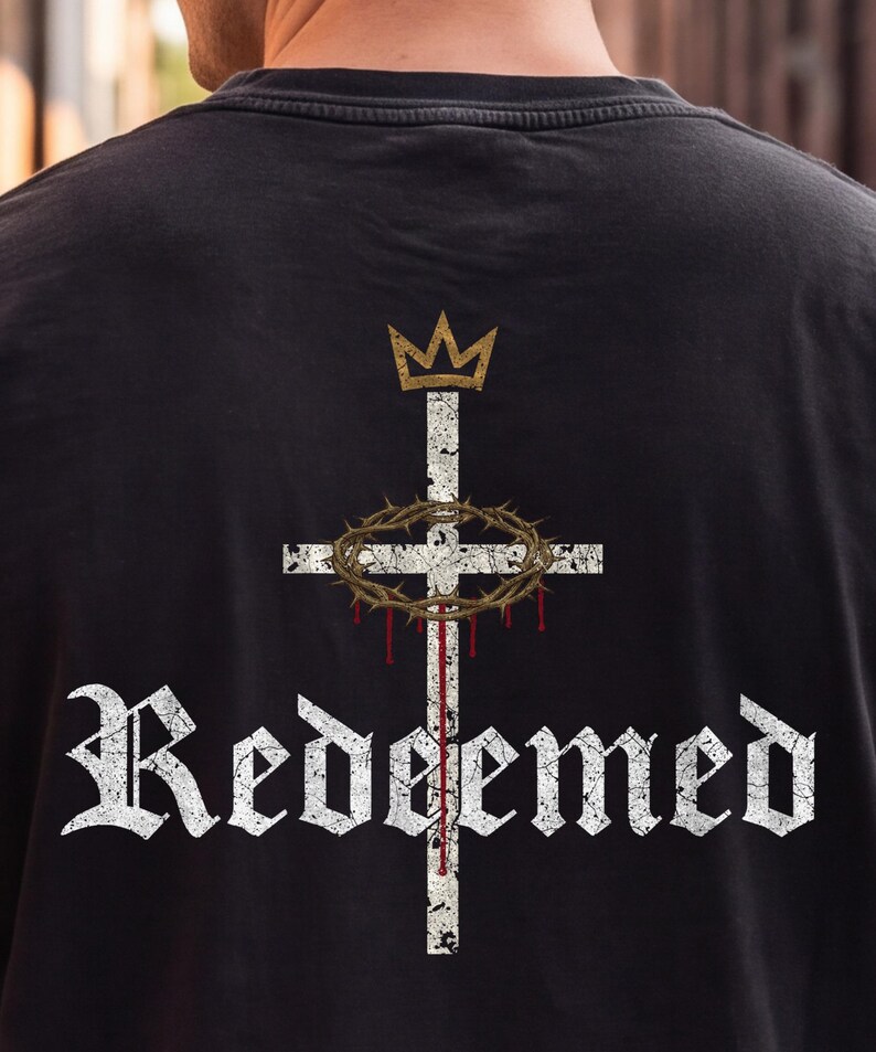 Op de afbeelding: Een zwart t-shirt met een afbeelding op de achterkant. Het ontwerp bevat een wit, verweerd kruis met een gouden kroon erboven, een bruine doornenkroon rond het kruis, rode druppels en het woord "Redeemed" in een wit gotisch lettertype.