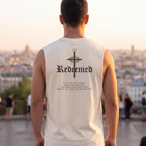 May include: A cream-coloured sleeveless shirt featuring a graphic on the back. The design includes a crown above a cross with a crown of thorns, the word "Redeemed" in black gothic font, and the text "If any man be in Christ, he is a new creature; old things are passed away; behold, all things are become new. 2 Corinthians 5:17."
