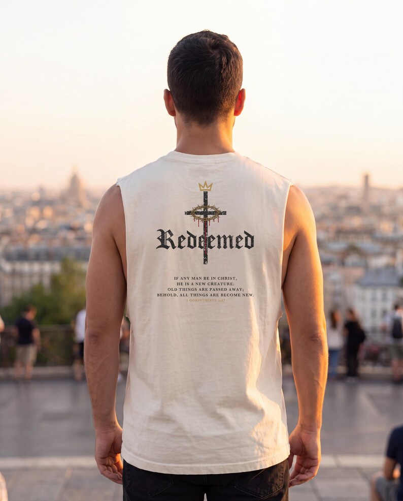 May include: A cream-coloured sleeveless shirt featuring a graphic on the back. The design includes a crown above a cross with a crown of thorns, the word "Redeemed" in black gothic font, and the text "If any man be in Christ, he is a new creature; old things are passed away; behold, all things are become new. 2 Corinthians 5:17."