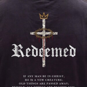 May include: A black t-shirt featuring a white distressed cross graphic with a gold crown above and a crown of thorns with red drips. The word "Redeemed" is printed in white gothic font, followed by the text "IF ANY MAN BE IN CHRIST, HE IS A NEW CREATURE: OLD THINGS ARE PASSED AWAY; BEHOLD, ALL THINGS ARE BECOME NEW. 2 CORINTHIANS 5:17".
