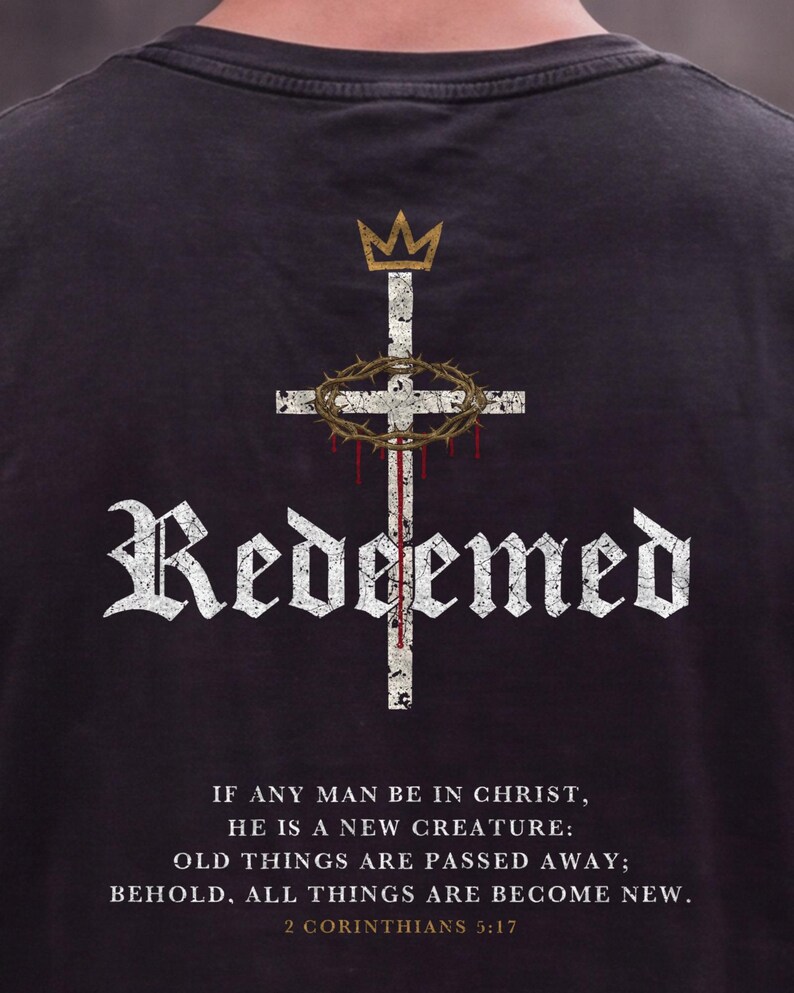 May include: A black t-shirt featuring a white distressed cross graphic with a gold crown above and a crown of thorns with red drips. The word "Redeemed" is printed in white gothic font, followed by the text "IF ANY MAN BE IN CHRIST, HE IS A NEW CREATURE: OLD THINGS ARE PASSED AWAY; BEHOLD, ALL THINGS ARE BECOME NEW. 2 CORINTHIANS 5:17".