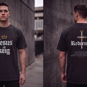 Op de afbeelding: Een zwart t-shirt met korte mouwen, voorzien van een gouden kroon en de witte tekst "Jesus is king" op de voorzijde. De achterkant toont een kruis met een doornenkroon, het woord "Redeemed" en een bijbelvers in witte tekst.