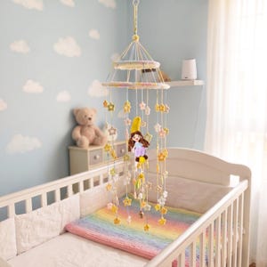 Op de afbeelding: Een hangende mobiel met een centrale gehaakte figuur die op een gele halve maan zit, omringd door meerdere strengen kleine, pastelkleurige gehaakte sterren die aan twee cirkelvormige, met garen omwikkelde ringen zijn opgehangen.
