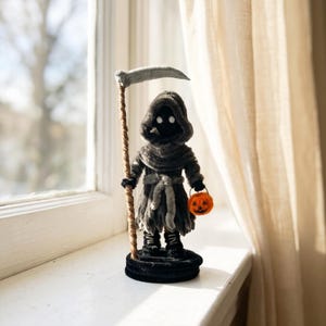Puede incluir: Una pequeña figura con capucha de color gris oscuro sobre una base circular negra. La figura viste una túnica harapienta con un fajín gris claro, botas negras y sostiene una guadaña alta con mango marrón texturizado y un pequeño cubo de calabaza naranja.