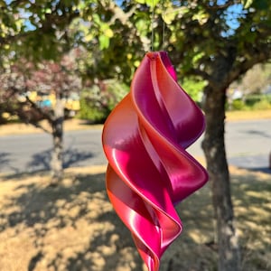 May include: A hanging spiral ornament featuring a smooth, curved, three-dimensional shape with a vibrant gradient colour transition from deep magenta and hot pink at the bottom to a bright orange and yellow hue at the top, suspended by a thin string.
