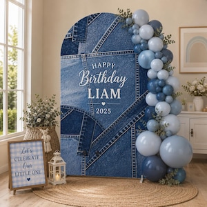Pode incluir: Um painel arqueado com estampa de patchwork de jeans azul com o texto HAPPY Birthday LIAM 2025, decorado com uma guirlanda vertical de balões azuis e cinzas e folhagem verde, posicionado sobre um tapete redondo trançado.