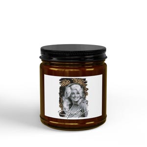 Puede incluir: Un frasco de vela de vidrio marrón con tapa negra de rosca. La etiqueta rectangular blanca presenta un retrato en blanco y negro de una persona sonriente con cabello rubio voluminoso, sobre un fondo de estampado de leopardo con el texto Dolly.