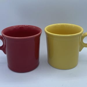 Könnte beinhalten: Zwei Keramikbecher mit einer glatten, glänzenden Oberfläche. Ein Becher ist tiefrot, der andere leuchtend gelb. Beide Becher haben einen zylindrischen Körper, einen breiten Rand und einen kreisförmigen Henkel an der Seite des Gefäßes.