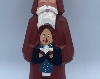 Op de afbeelding: Een handgesneden houten beeldje van een lang figuur in een lange, donkerrode mantel met capuchon, witte baard en snor, die een kleinere pop vasthoudt met bruin haar, een blauwe jurk met witte stippen en een zwart jasje aan de voorzijde.