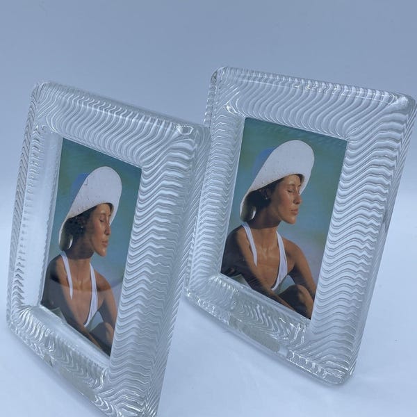 May include: Two rectangular picture frames made of clear glass with a textured, wavy, rippled pattern along the borders. Each frame contains a photograph of a person wearing a white hat and white top, positioned against a soft, light-colored background.