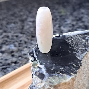 May include: A white oval-shaped bead with a smooth, polished surface. The bead has a subtle, iridescent sheen and is made of natural material.