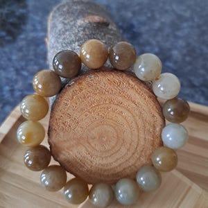 May include: A beaded bracelet featuring polished, round beads in shades of brown, tan, and cream. The bracelet is displayed on a wooden slice, showcasing the natural variations in color and texture of the beads. The bracelet is a jewelry item.