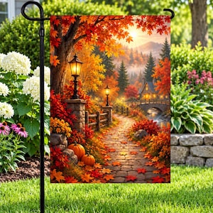May include: A rectangular garden flag featuring a vibrant autumn landscape with a stone path, two glowing black street lamps, orange pumpkins, and a small house near a bridge, all surrounded by trees with red, orange, and yellow fall foliage.