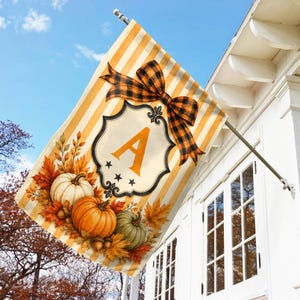 May include: A decorative garden flag featuring a vertical yellow and white striped background. The design includes a central ornate frame with the letter A, a large orange and black plaid bow, and a cluster of pumpkins and autumn leaves.