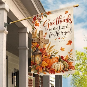May include: A rectangular garden flag featuring the text "Give thanks to the Lord, for He is good. PSALM 107:1" in brown script. The design includes a wooden cross, a lit lantern, pumpkins, wheat stalks, red berries, and autumn leaves in orange and yellow.