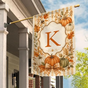May include: A vertical garden flag featuring a cream and beige striped background with a central scalloped frame containing the letter K. The design is decorated with orange pumpkins, white and green gourds, autumn leaves, and a plaid bow.
