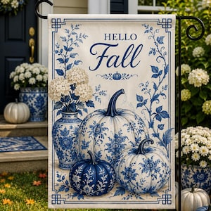 May include: A rectangular garden flag featuring a white background with blue floral patterns. The text HELLO Fall is printed at the top. The design includes a vase filled with white flowers and two pumpkins decorated with blue and white floral motifs.